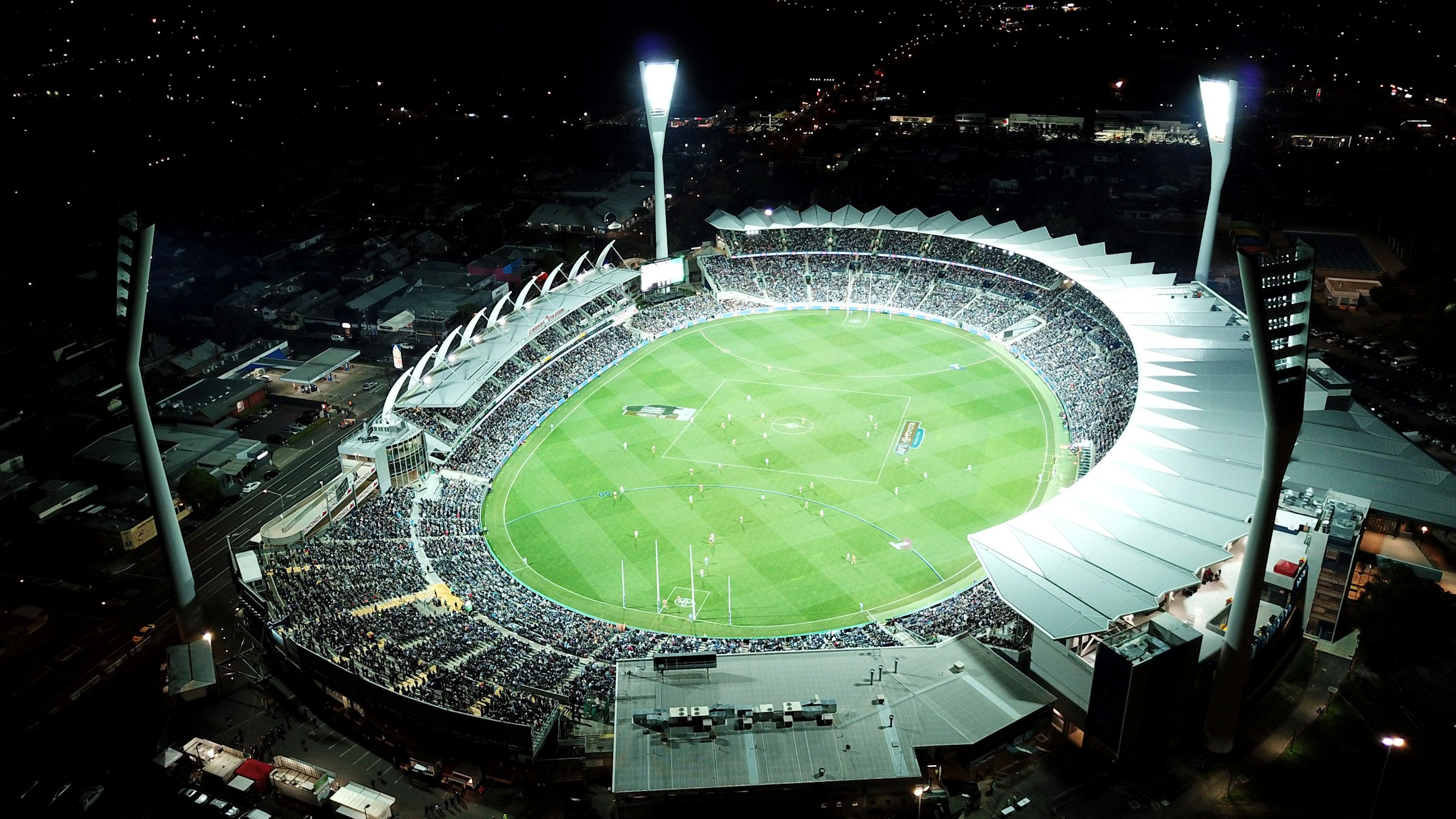 A floodlit Australian rules football match at night, seen from above, with a full stadium crowd